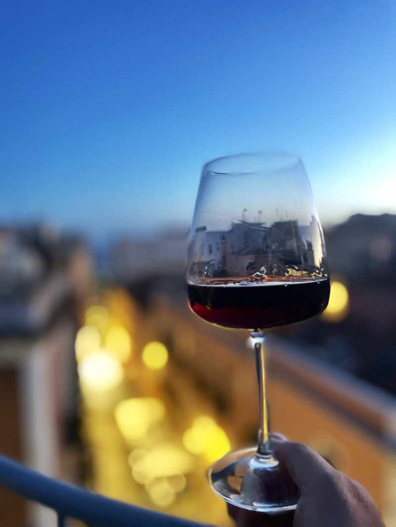 POV hand holding a wine glass at its base overlooking the Marina district of Cagliari at sunset with festively lit street below out-of-focus in the background.